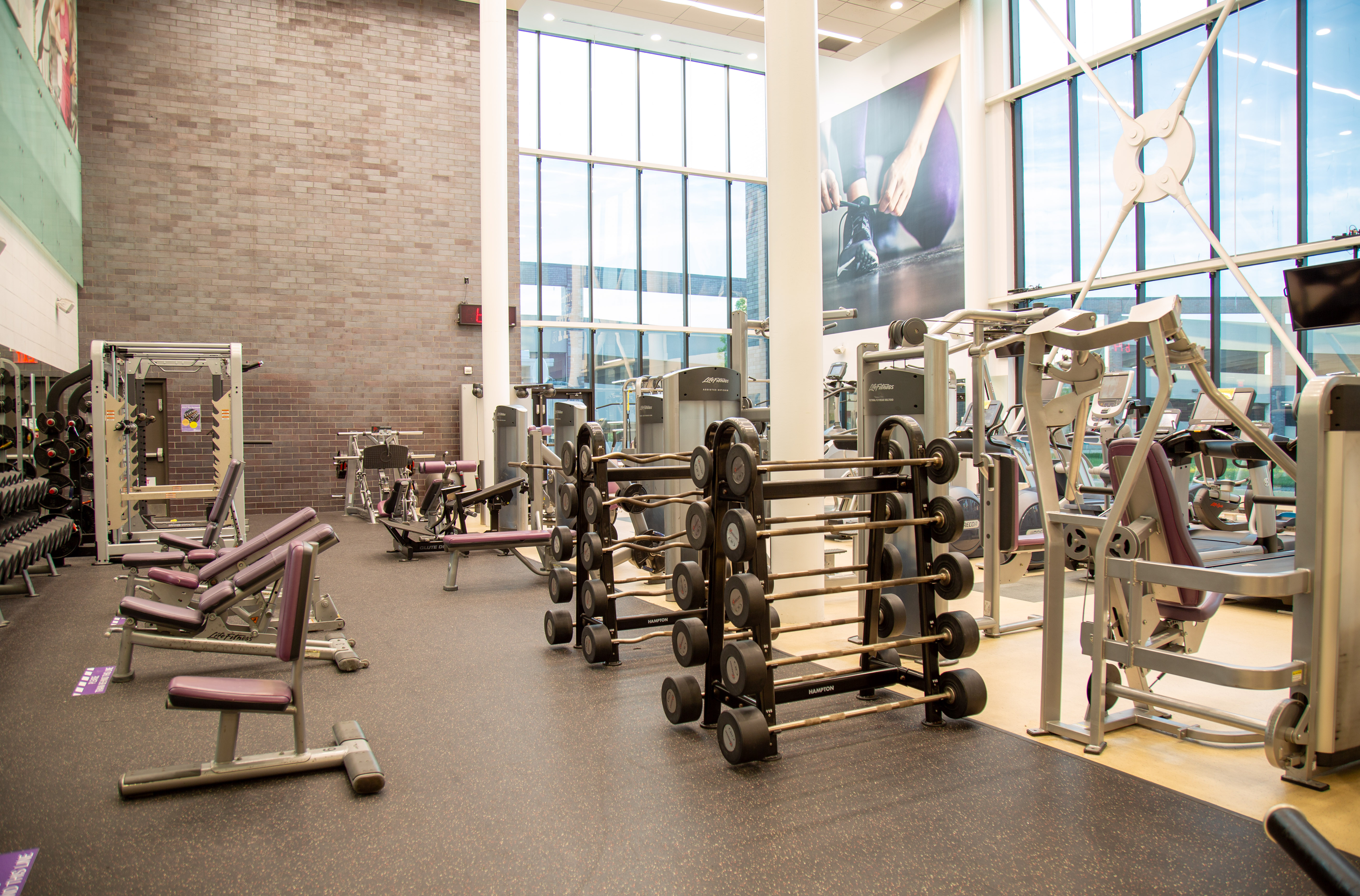 Spacious gym area with weight racks, benches, and various strength training machines under large windows.