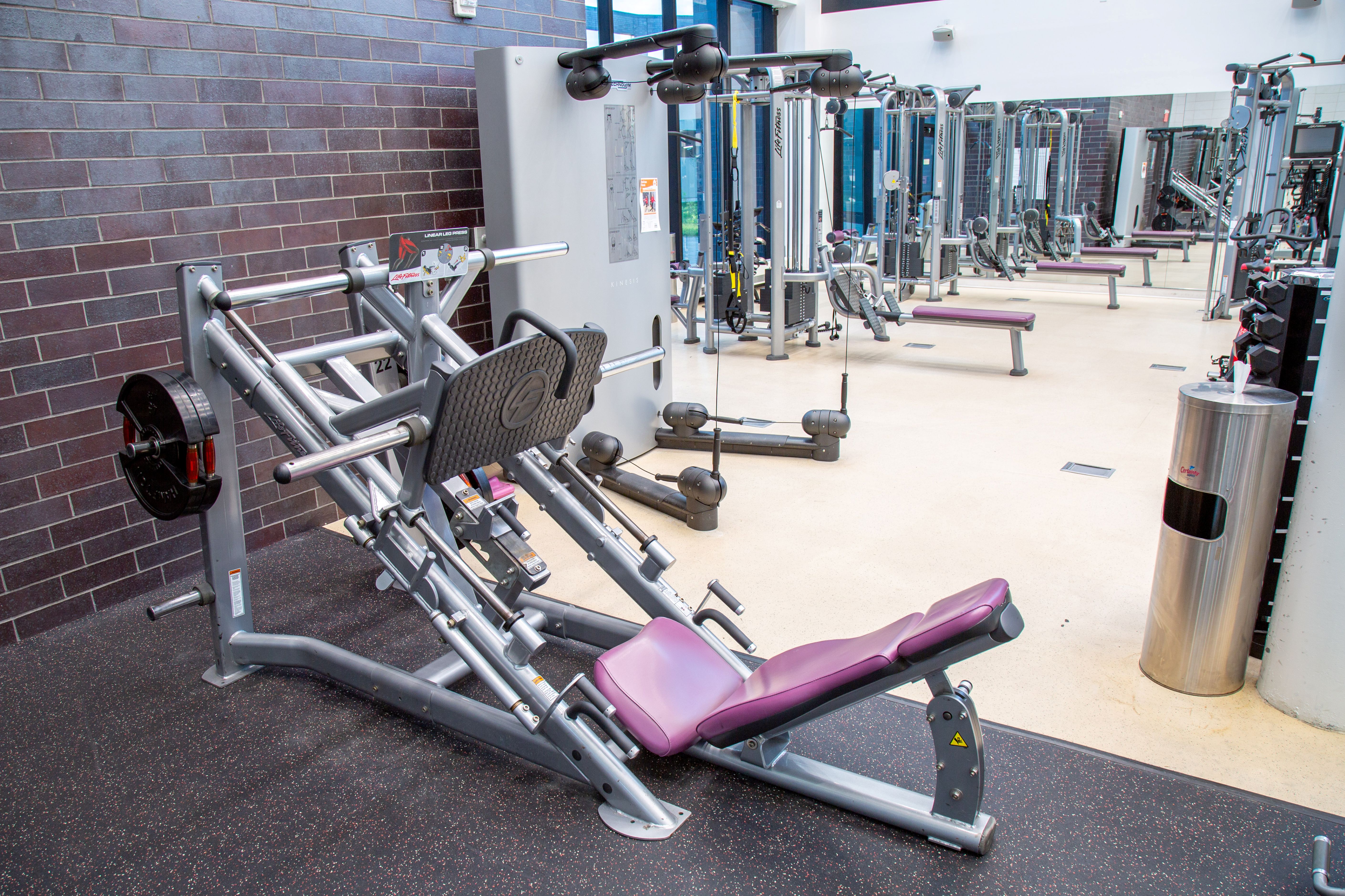 Modern gym interior featuring a leg press machine with purple padding and various strength training equipment in the background.