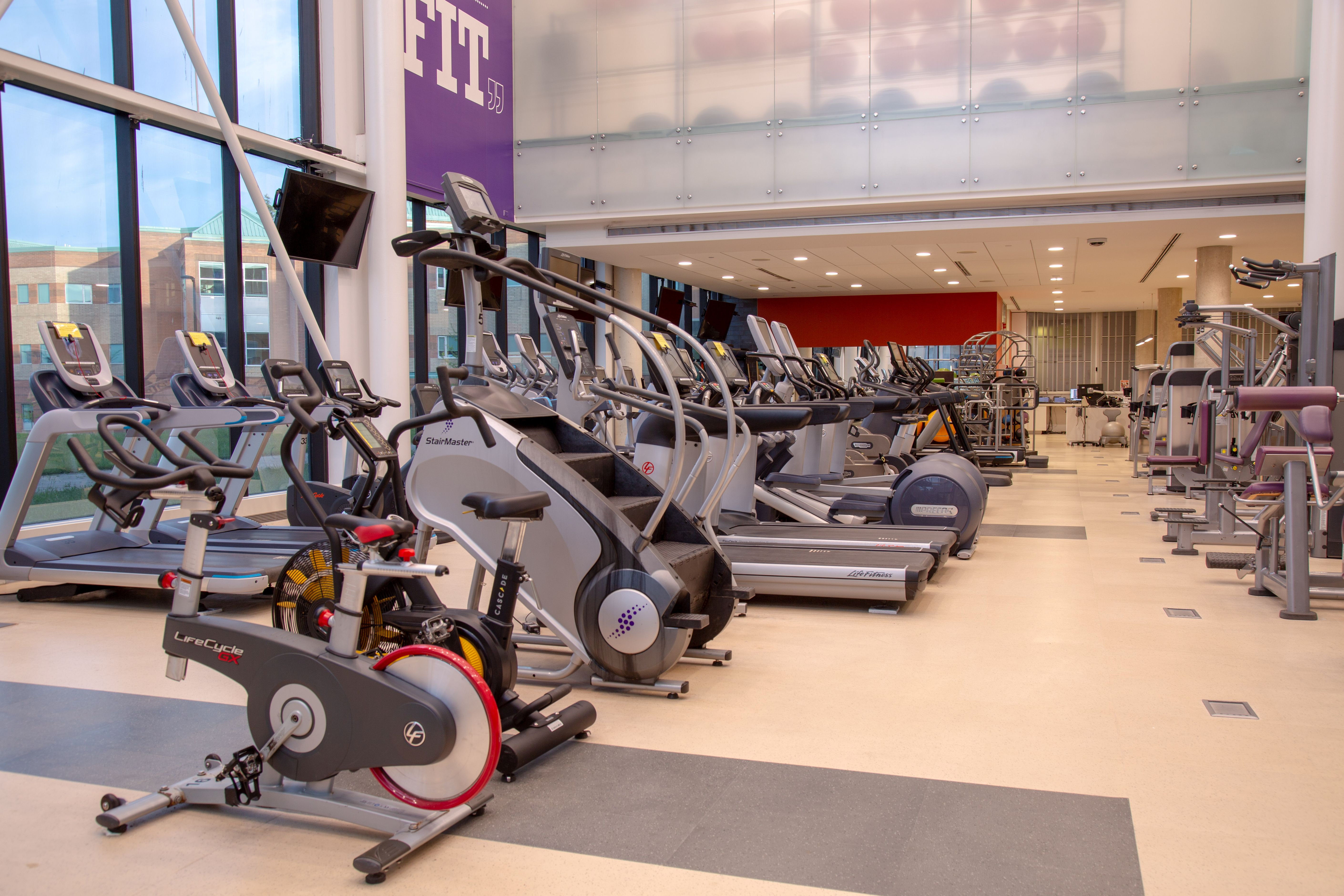 Modern gym interior with rows of treadmills, stationary bikes, and elliptical machines under bright lighting.