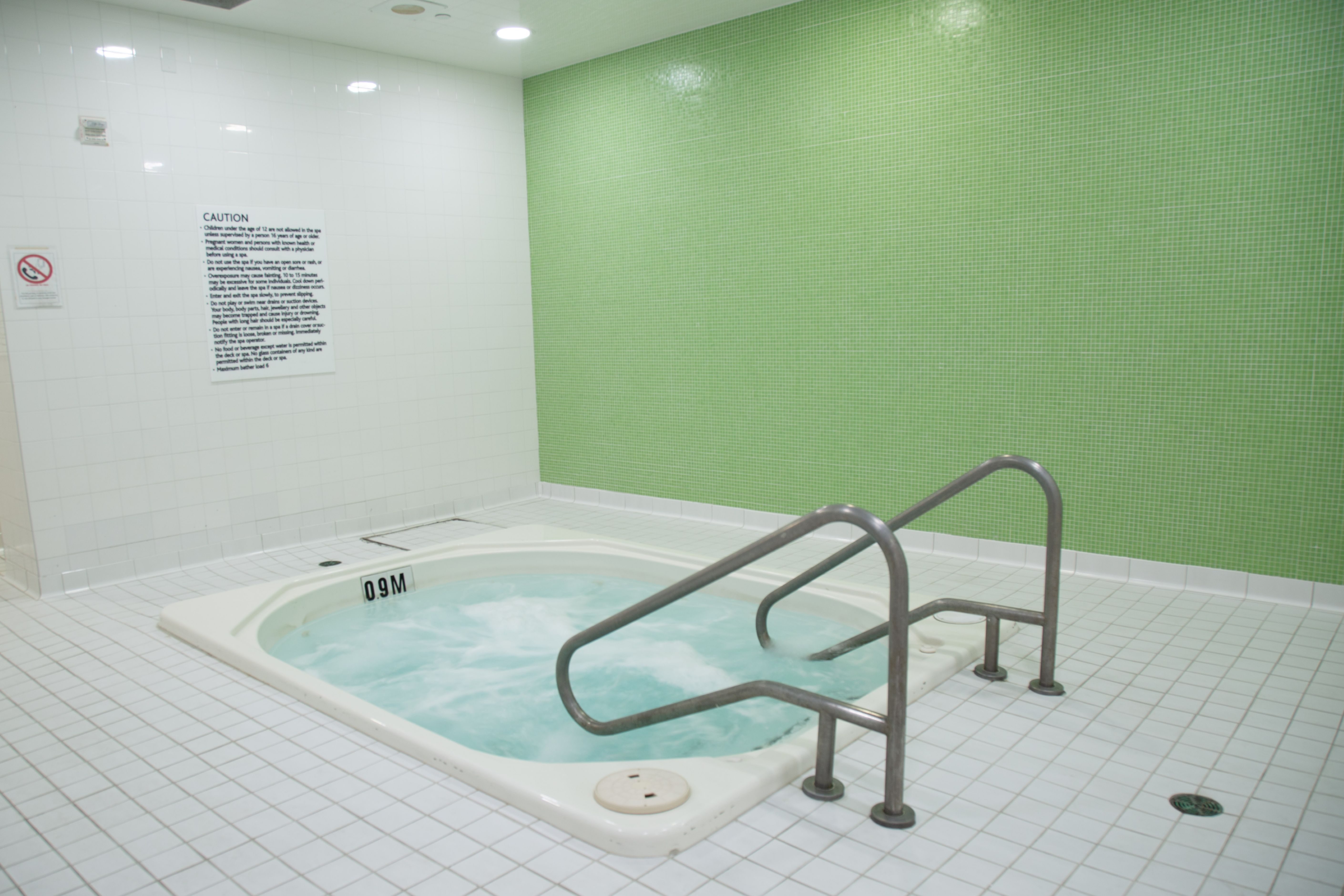 Indoor hot tub with bubbling water, surrounded by white tiled floor and green tiled wall, featuring metal handrails for safe entry.