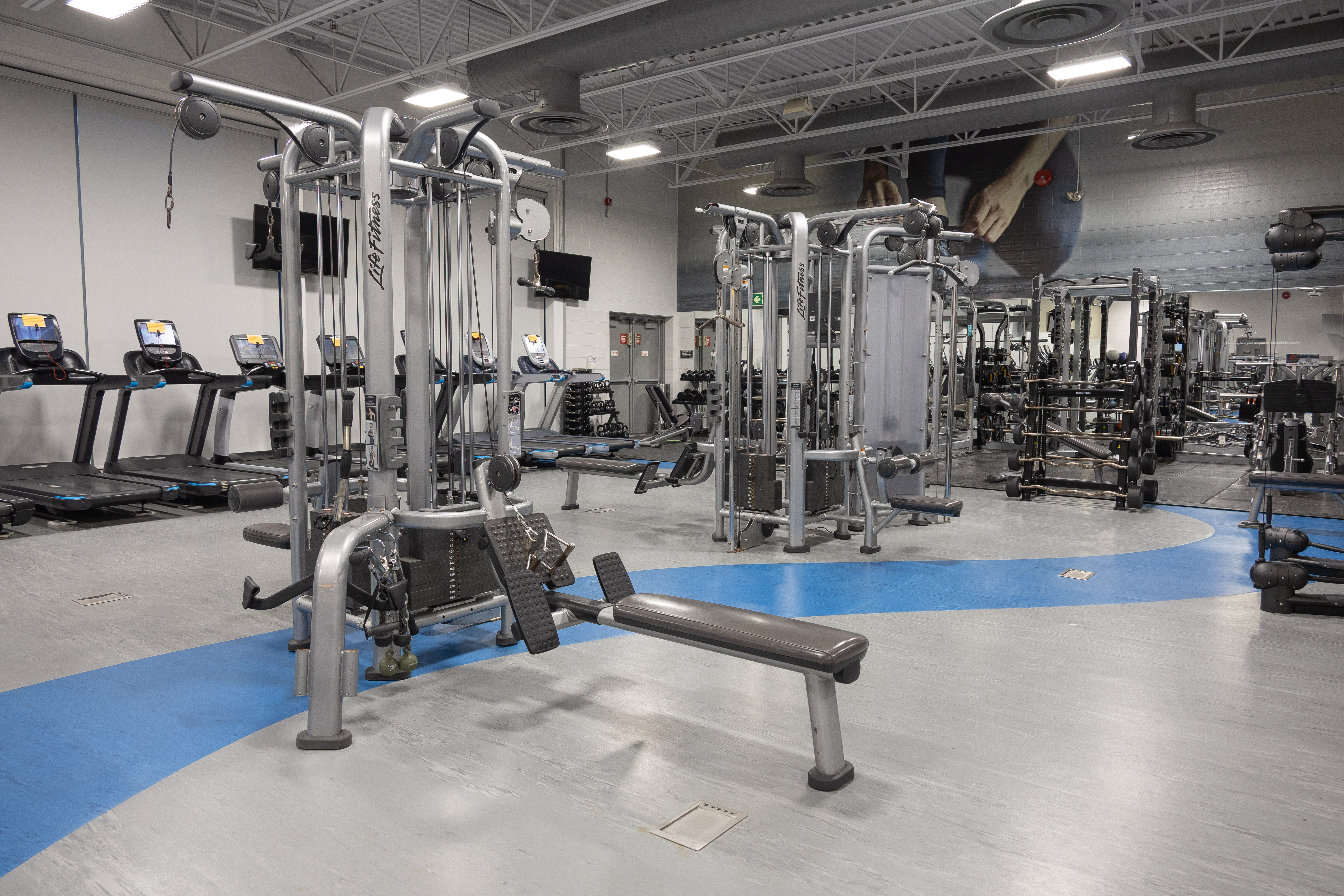 Spacious gym interior with weight machines, treadmills, and mirrored walls under bright overhead lighting.