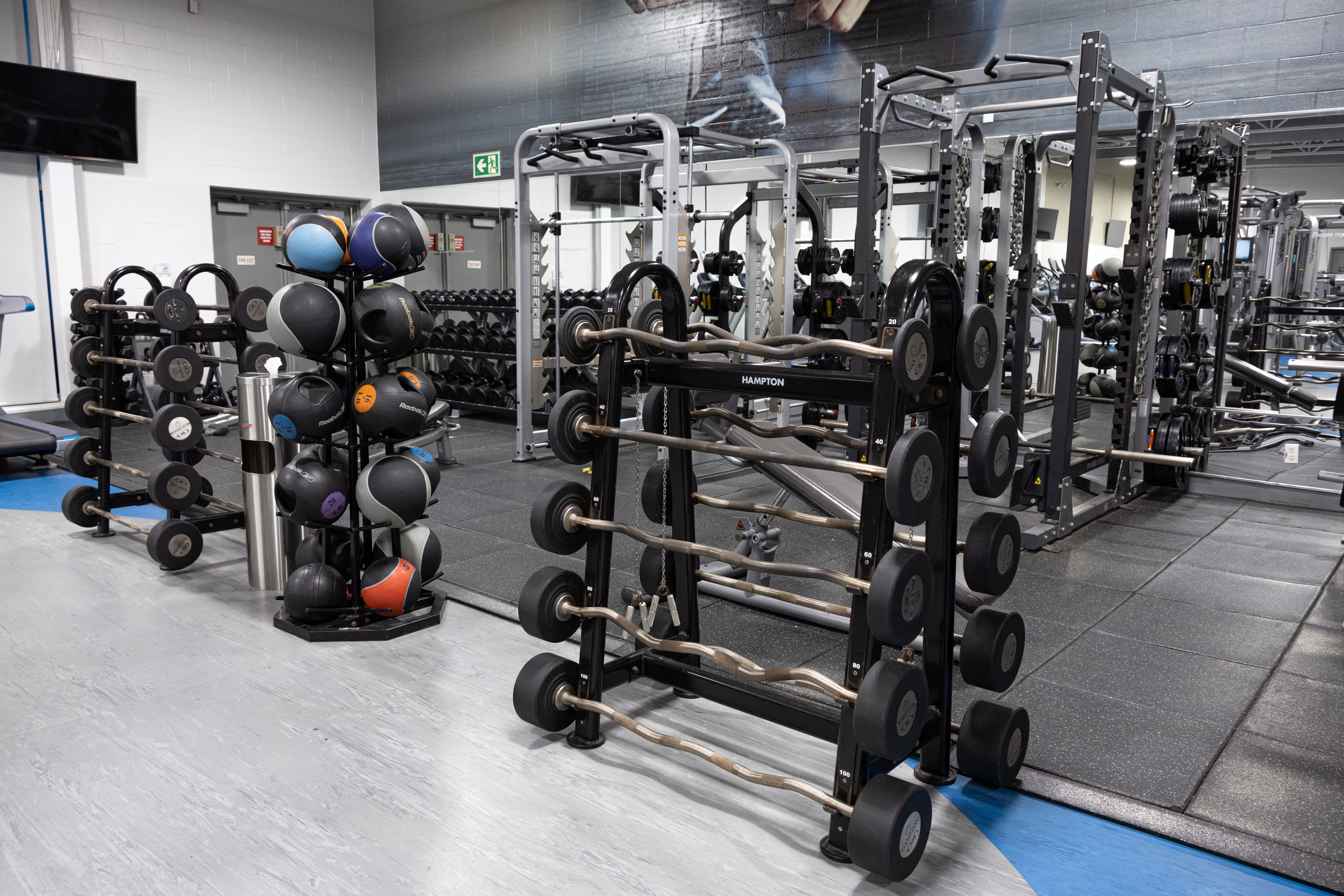 Modern gym area with racks of barbells, medicine balls, and dumbbells neatly organized against mirrored walls.