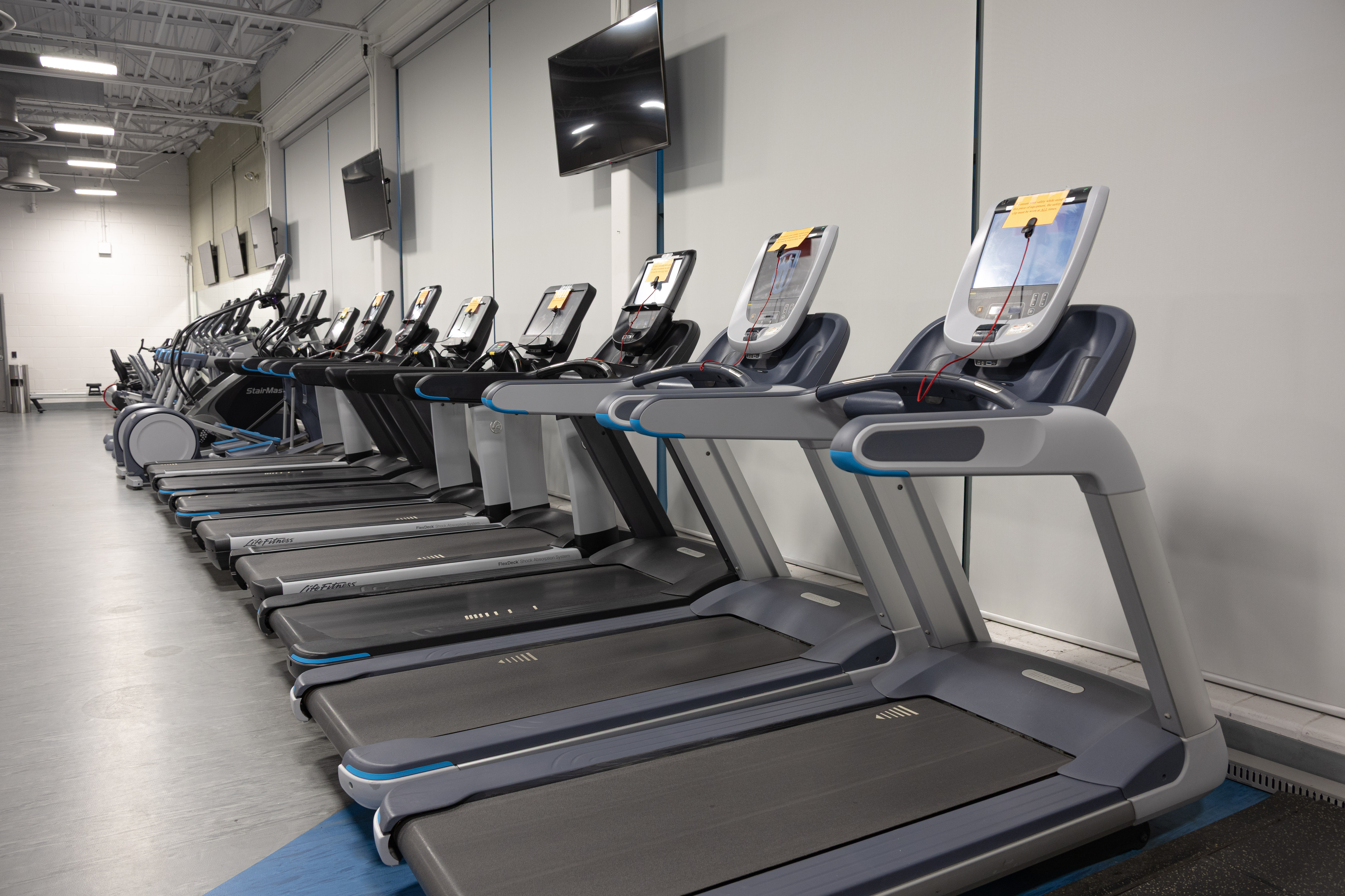 A row of modern treadmills lined up in a spacious, well-lit gym with large screens mounted on the wall above them.