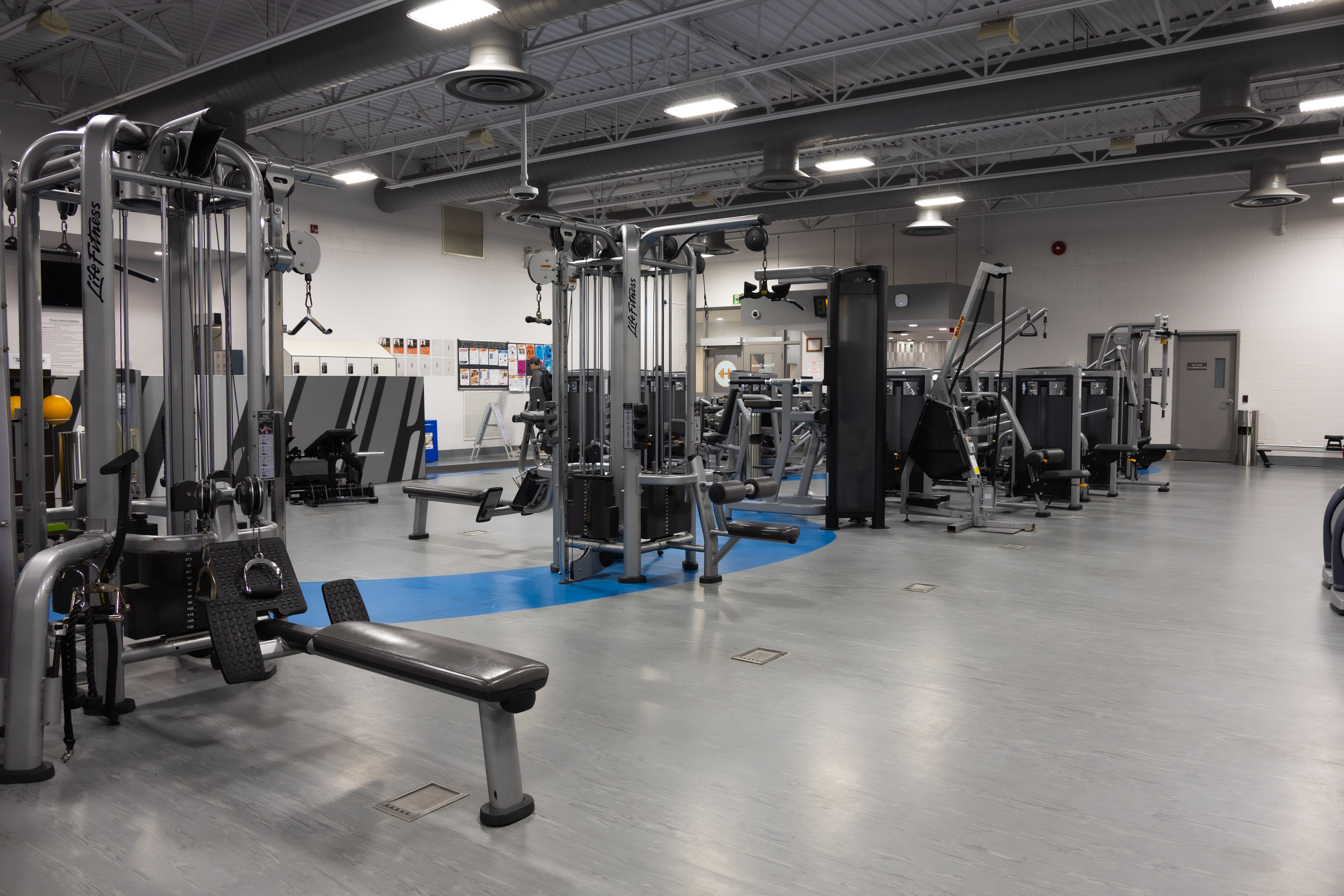 Spacious gym interior with various strength training machines and benches arranged on a clean gray floor under bright overhead lights.