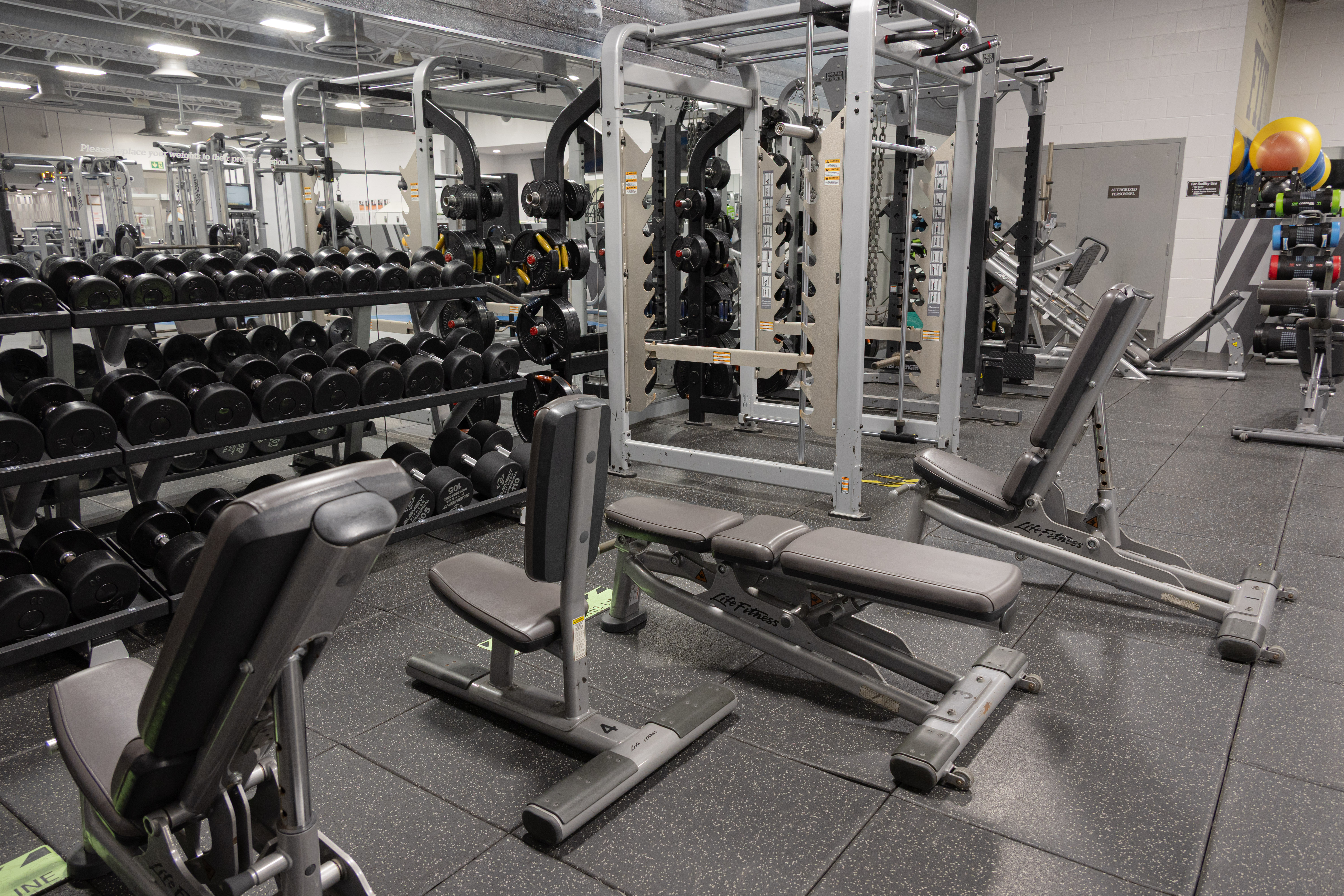 Empty gym area with weight benches, dumbbell racks, and various strength training machines under bright overhead lights.