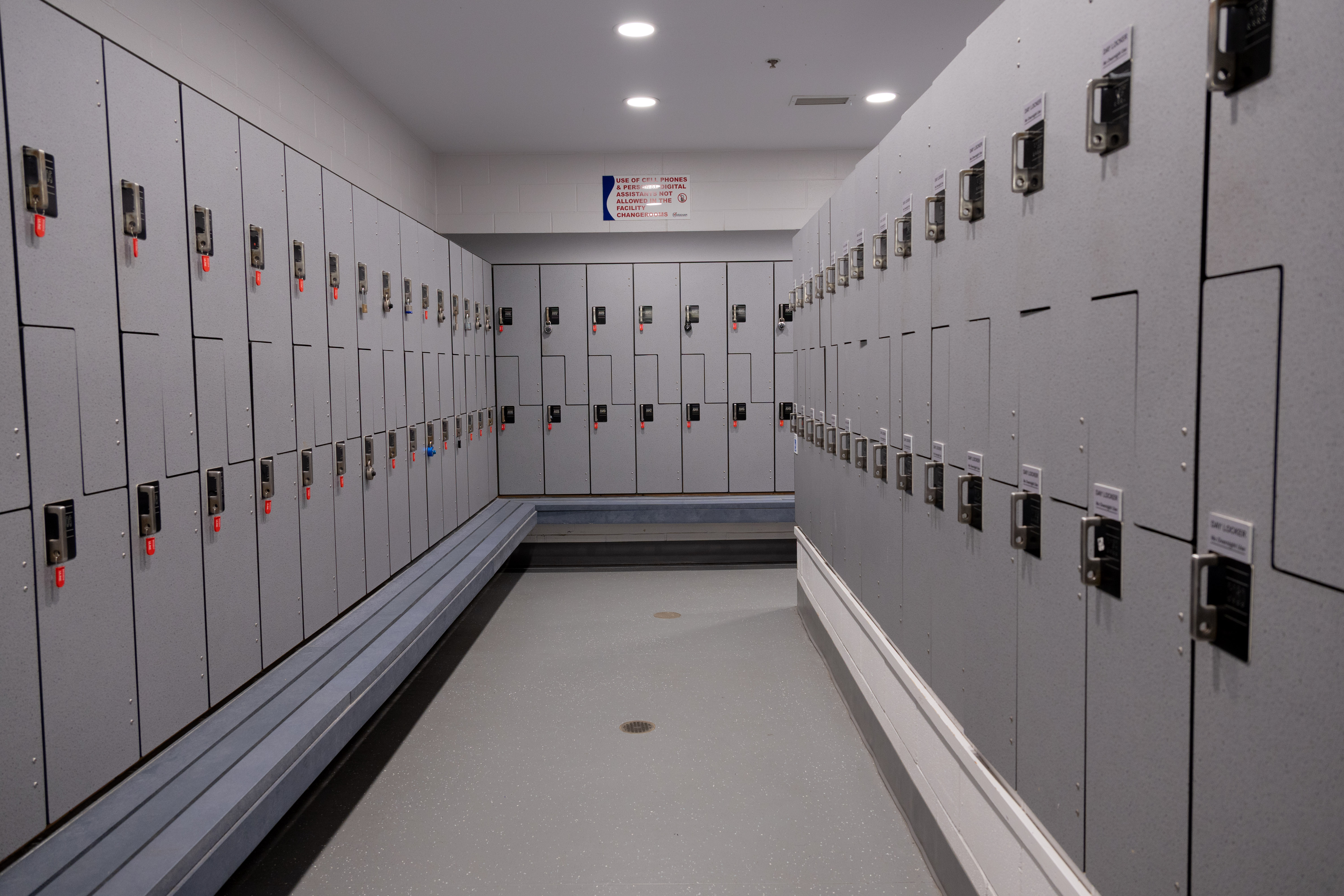 Long corridor of gray lockers with combination locks and a bench along one side in a well-lit locker room.