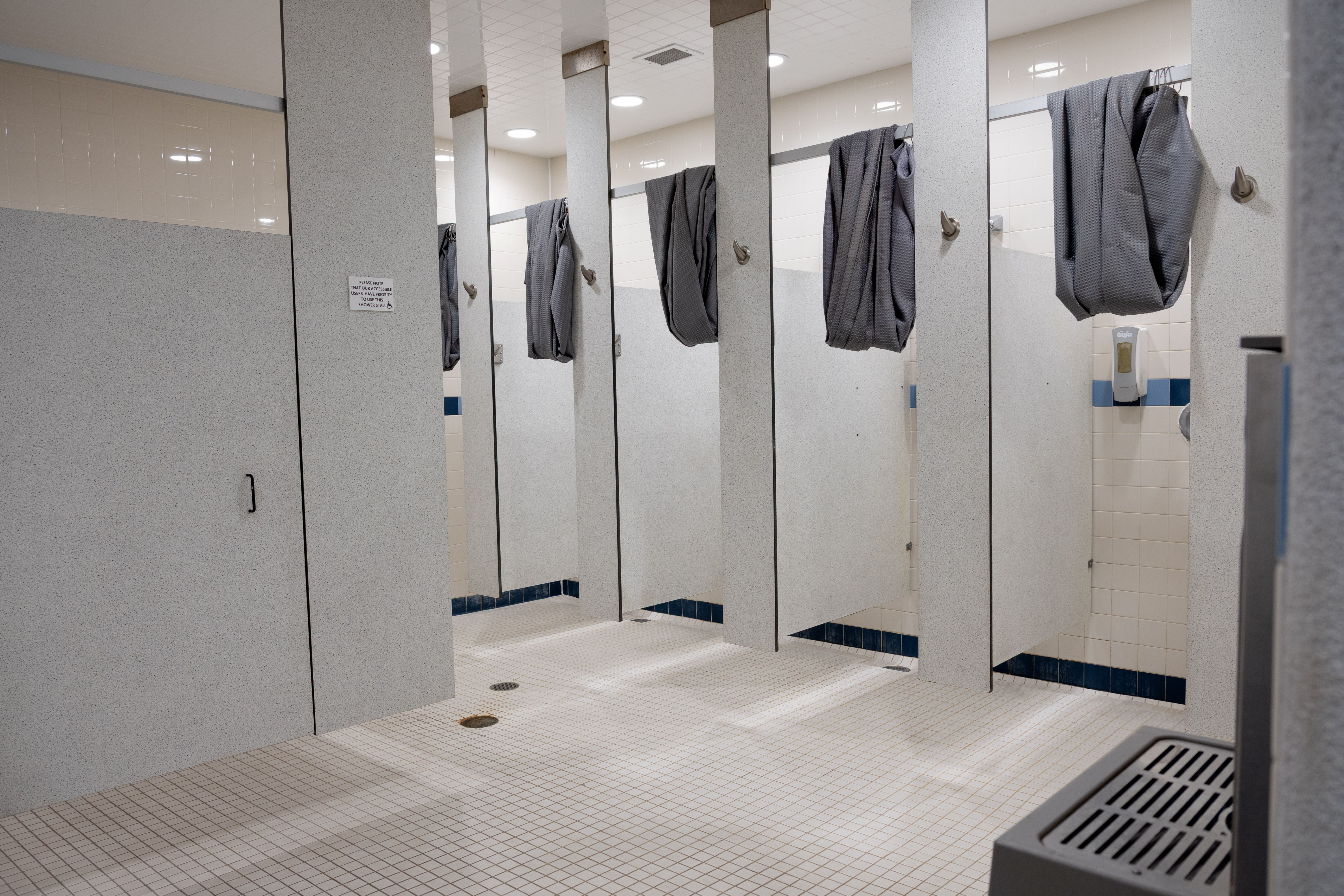 Locker room shower area with multiple stalls, each featuring a gray towel hanging on the door hooks and tiled floors and walls.