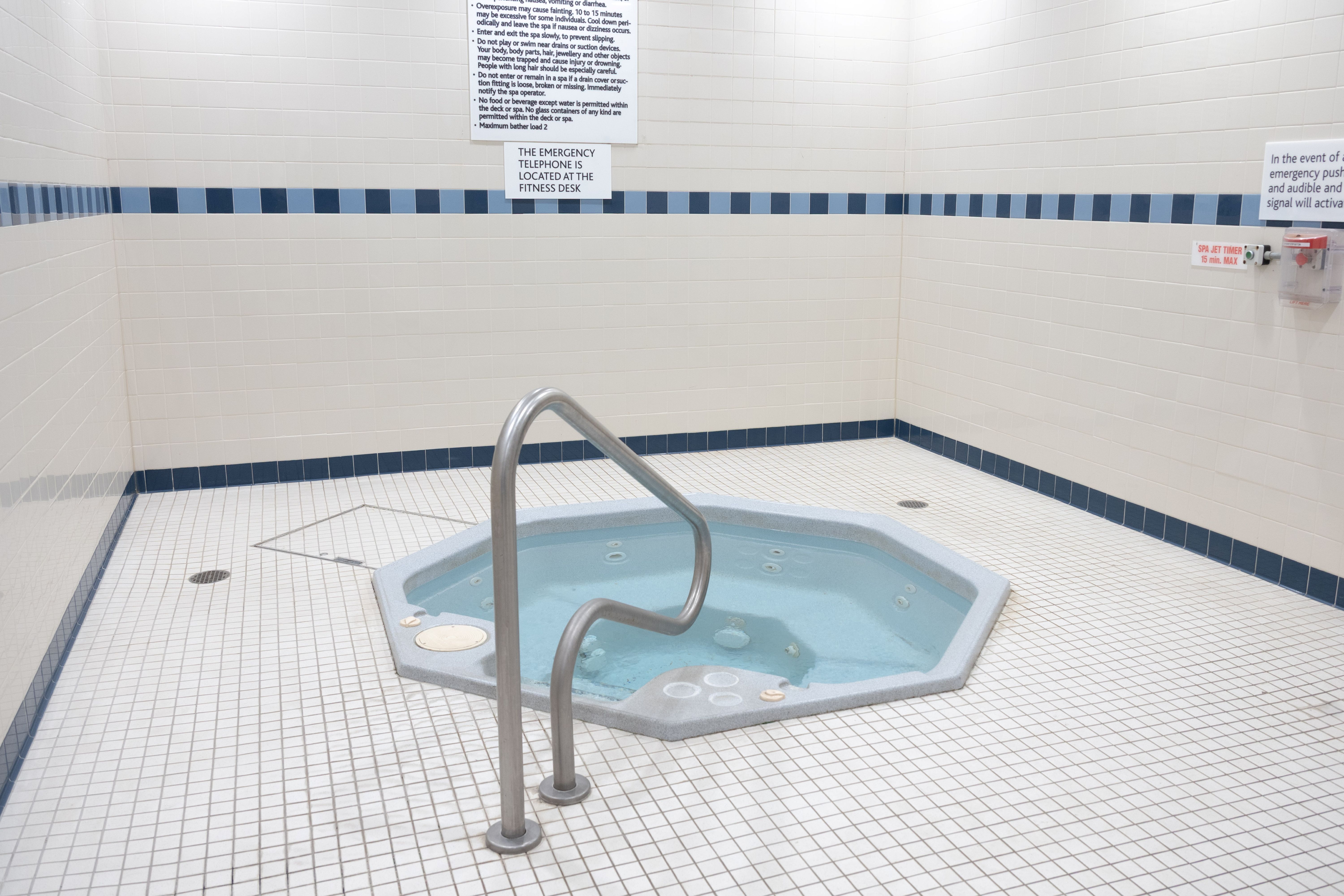 Empty indoor hot tub with clear water and stainless steel handrail surrounded by white tiled walls and floor.