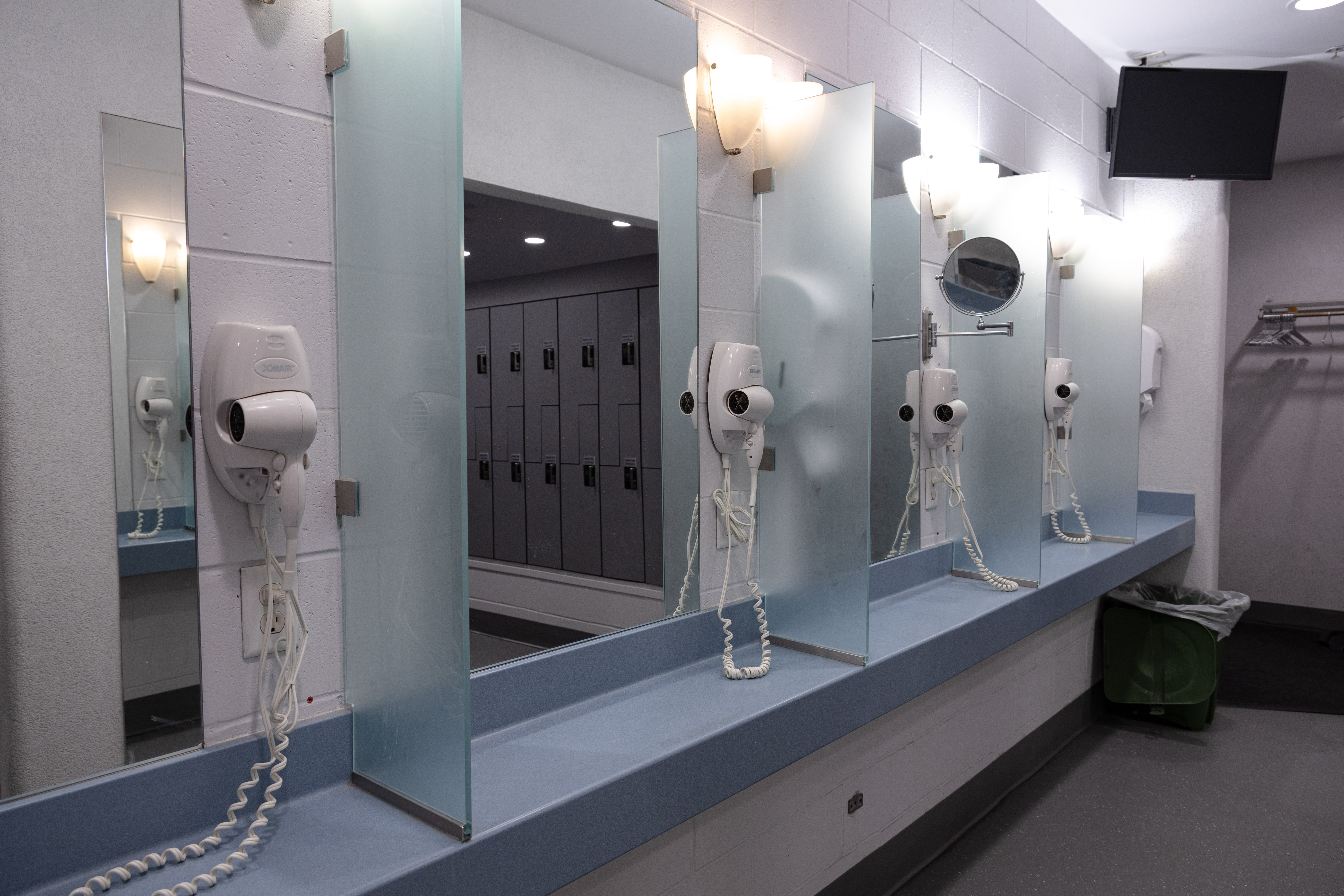 Row of mirrors with wall-mounted hair dryers above a long blue countertop in a locker room bathroom.