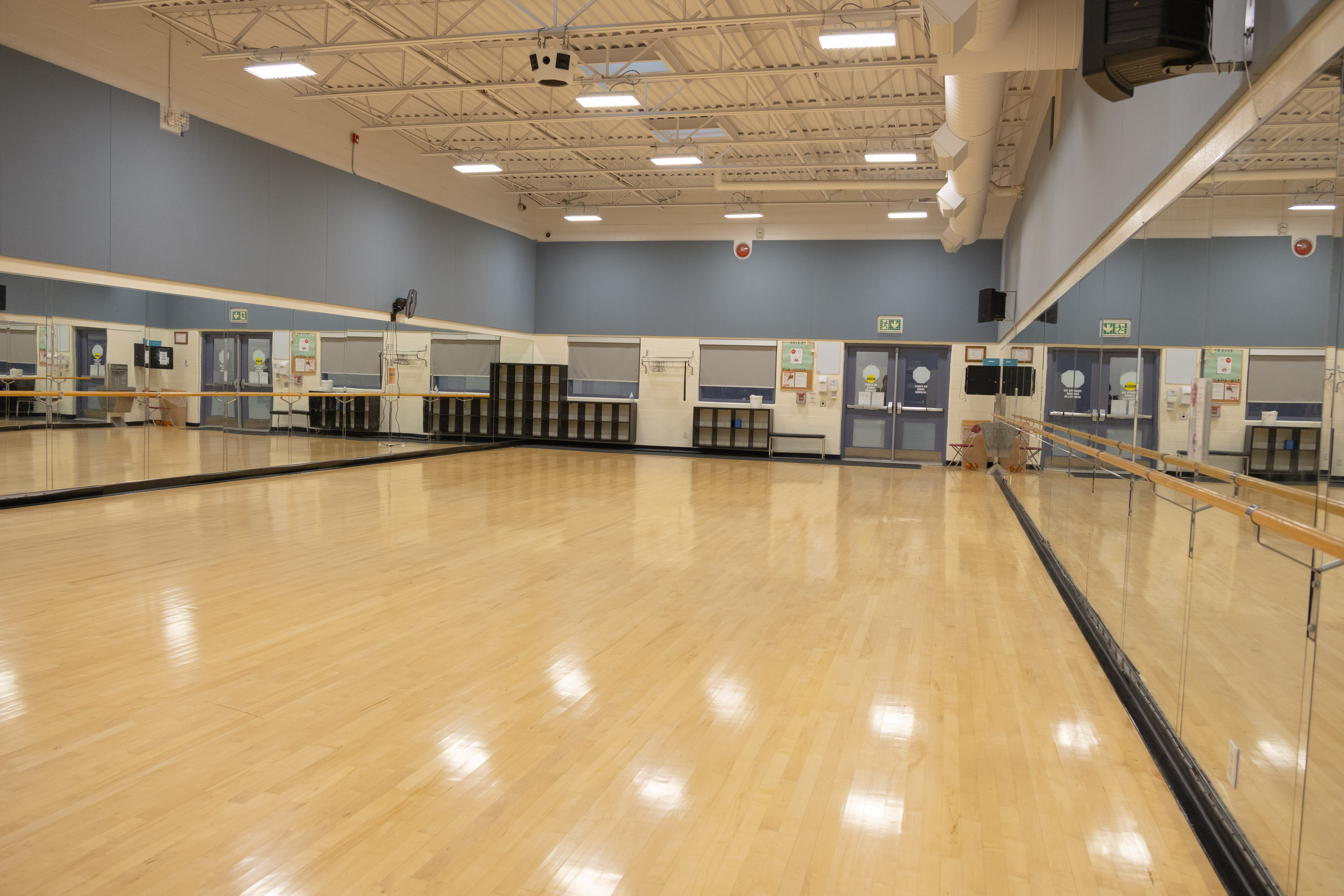 Spacious dance studio with polished wooden floors, mirrored walls, and ballet barres under bright ceiling lights.
