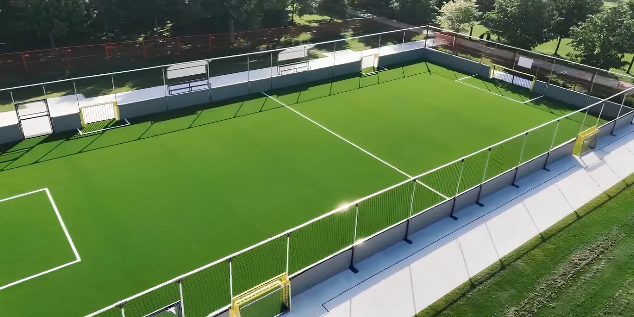 A fenced outdoor artificial turf soccer field viewed from above, with white field markings and surrounding greenery.