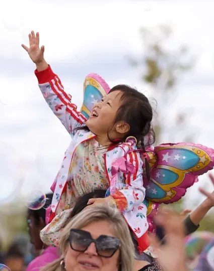Child with butterfly wings sitting on parent's shoulders reaches for bubbles in the air