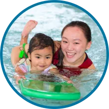 Swim instructor smiling at the camera with a child swimming with a float board