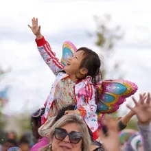Child with butterfly wings sitting on parent's shoulders reaches for bubbles in the air