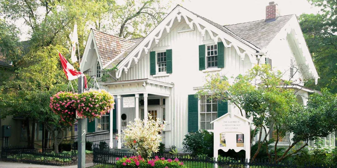 Exterior view of the McKay Art Centre, a historic white heritage house with green trim, decorative gables, and a covered porch, surrounded by mature trees and flowering gardens. A Canadian flag and hanging flower baskets are visible near the entrance.