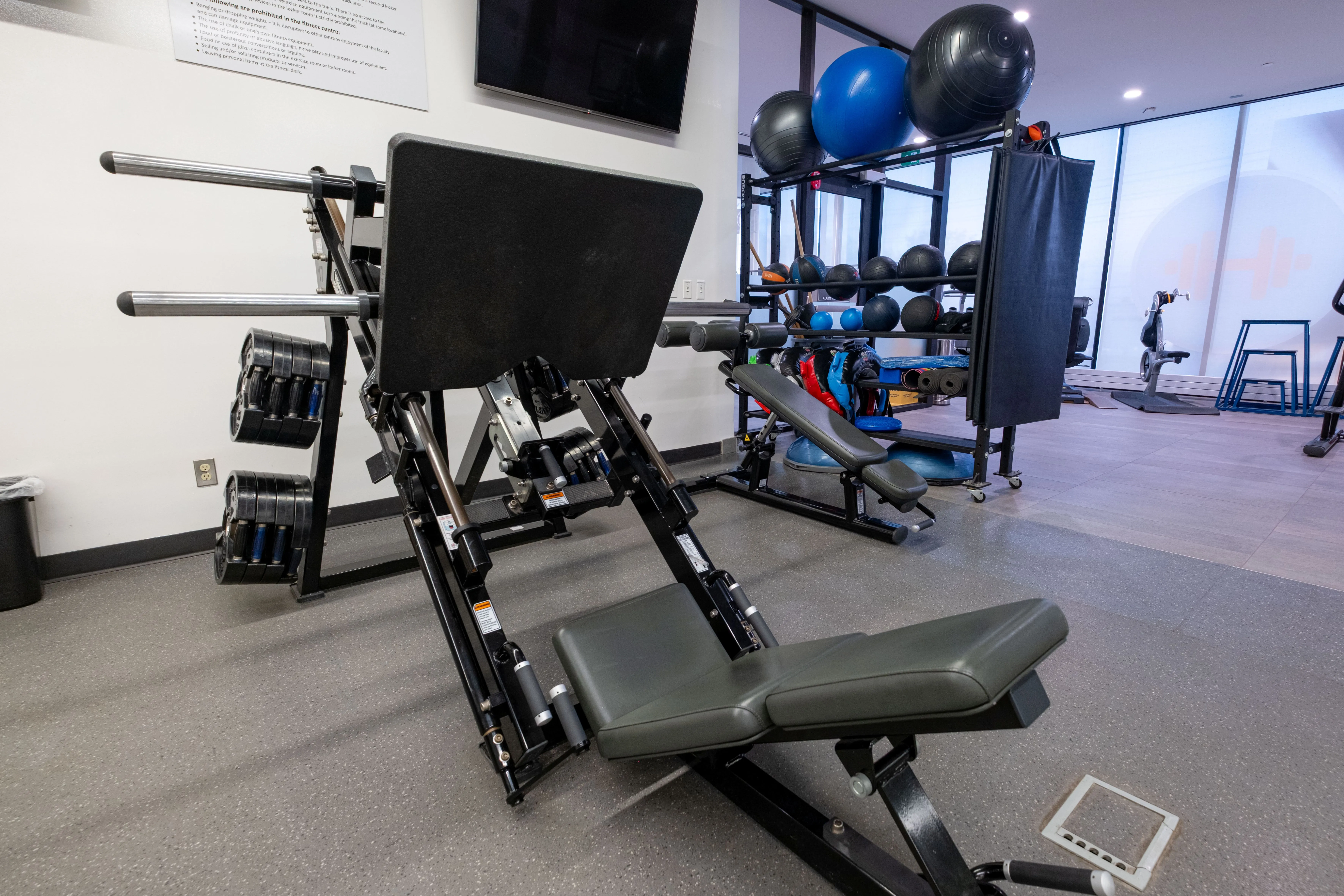 Leg press machine with padded seat and footplate in a modern gym setting with exercise balls and weights in the background.