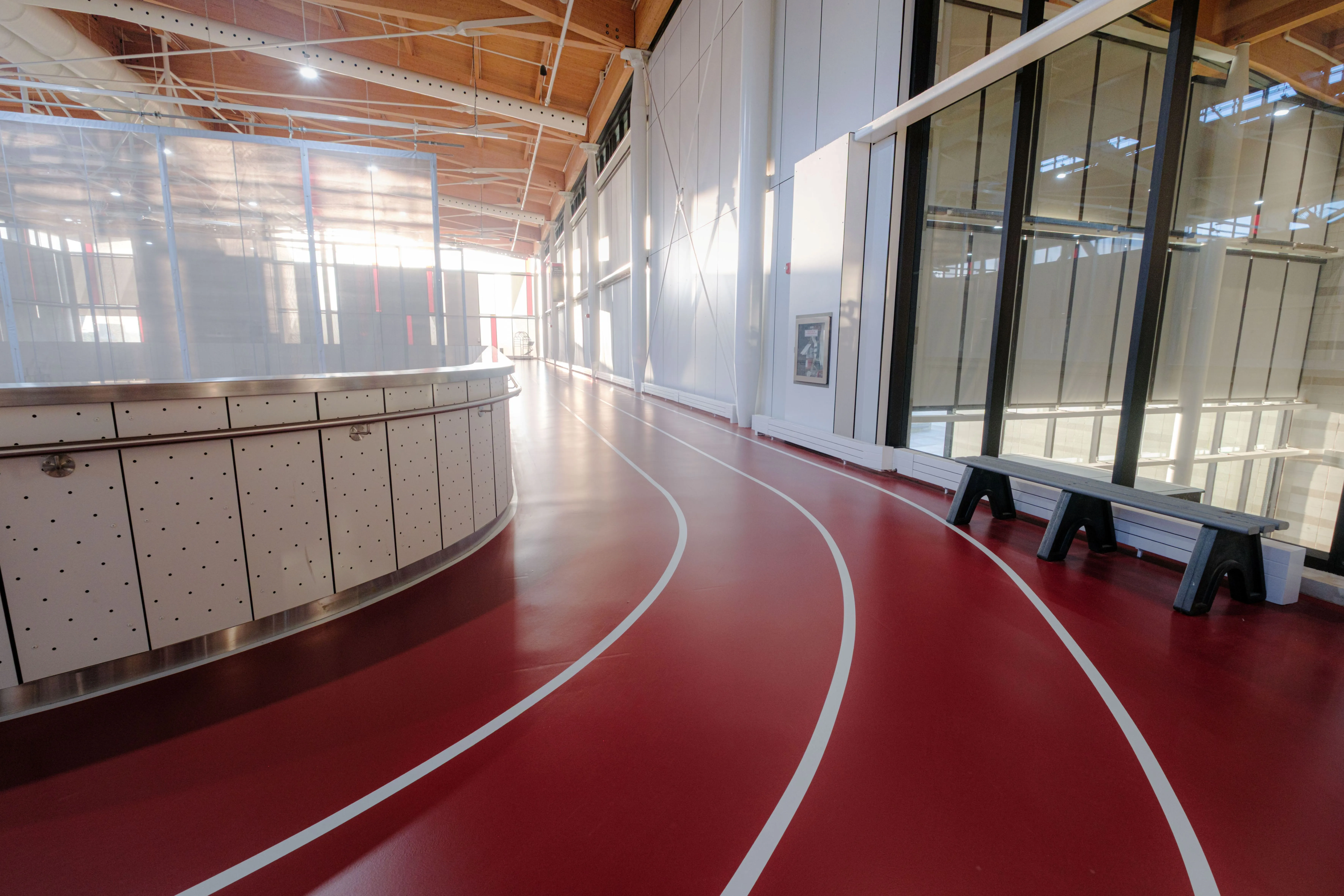 Indoor running track with red surface and white lane markings inside a spacious gym with large windows and wooden ceiling beams.