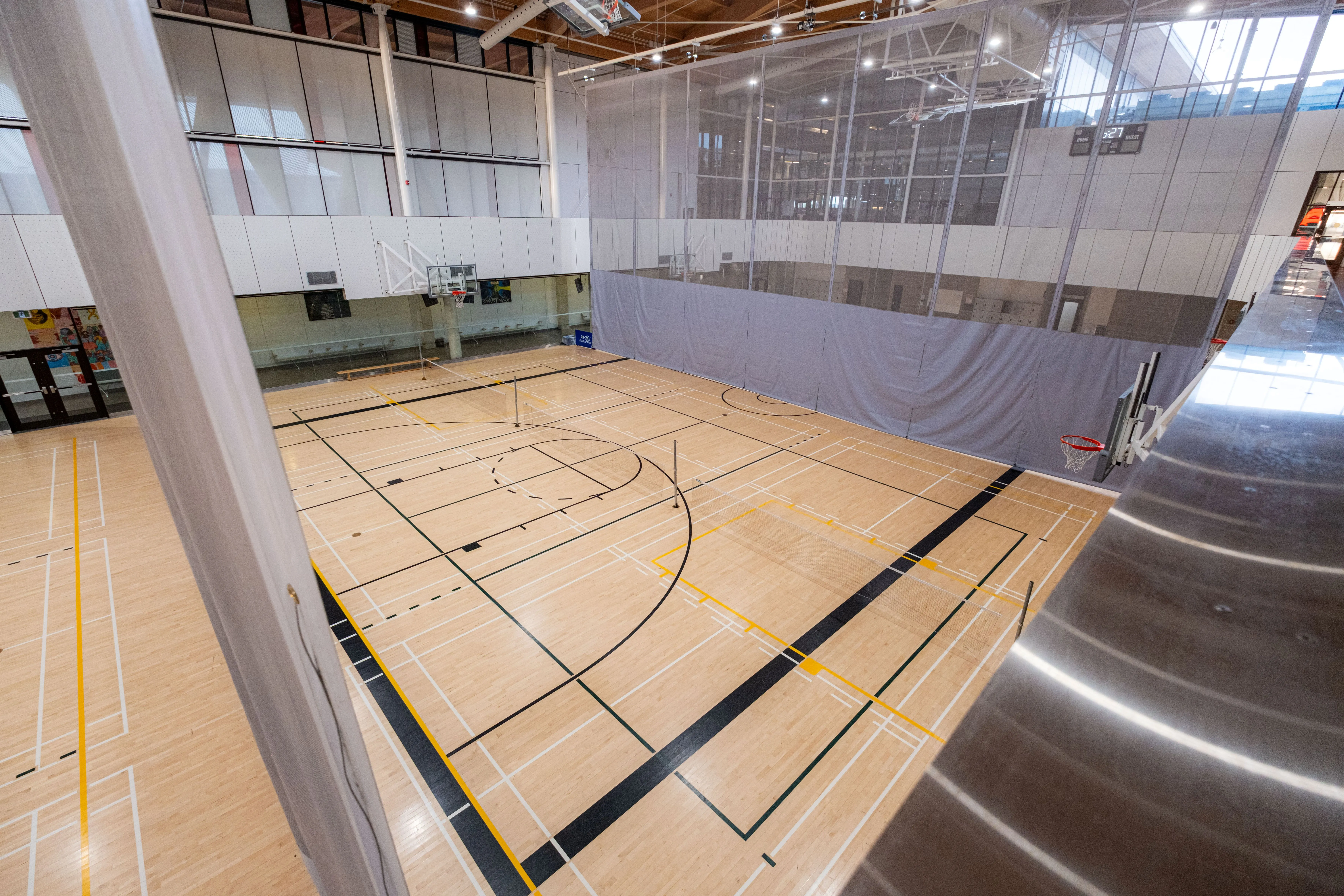 Empty indoor basketball court with polished wooden floor and multiple sports markings under bright overhead lights.