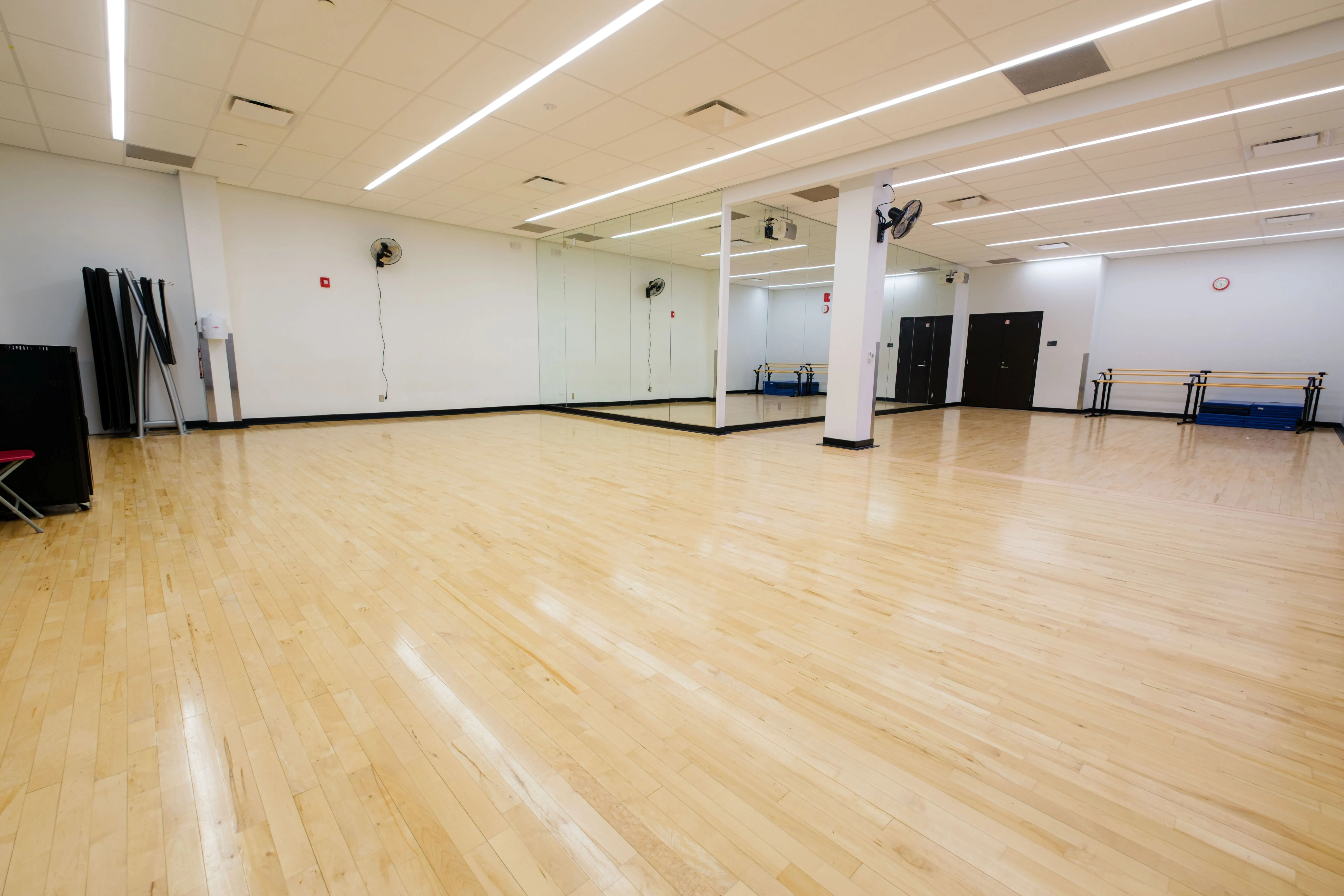 Spacious empty dance studio with polished wooden floors and large wall mirrors under bright ceiling lights.