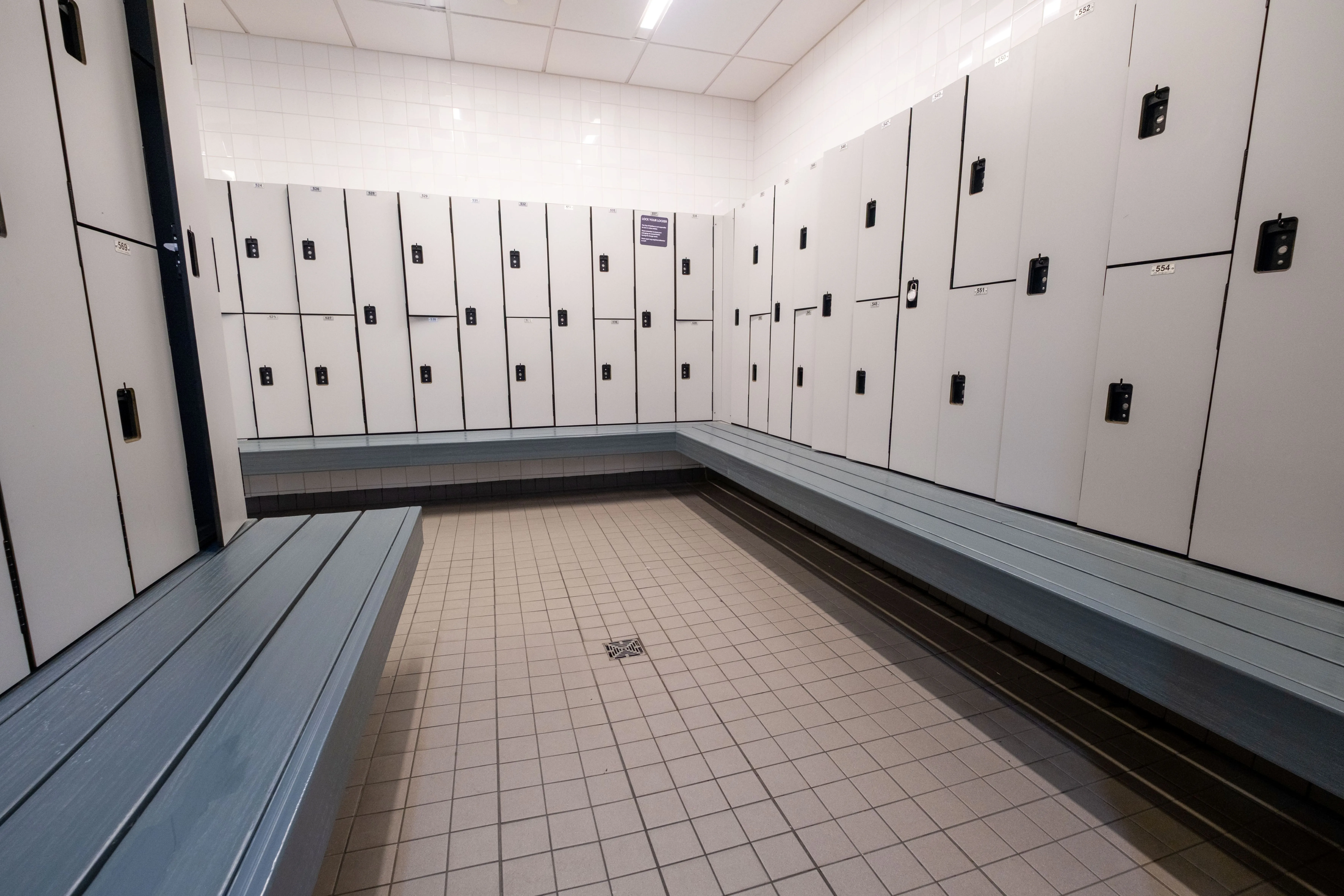 Empty locker room with white lockers lining the walls and blue benches along the perimeter on tiled flooring.