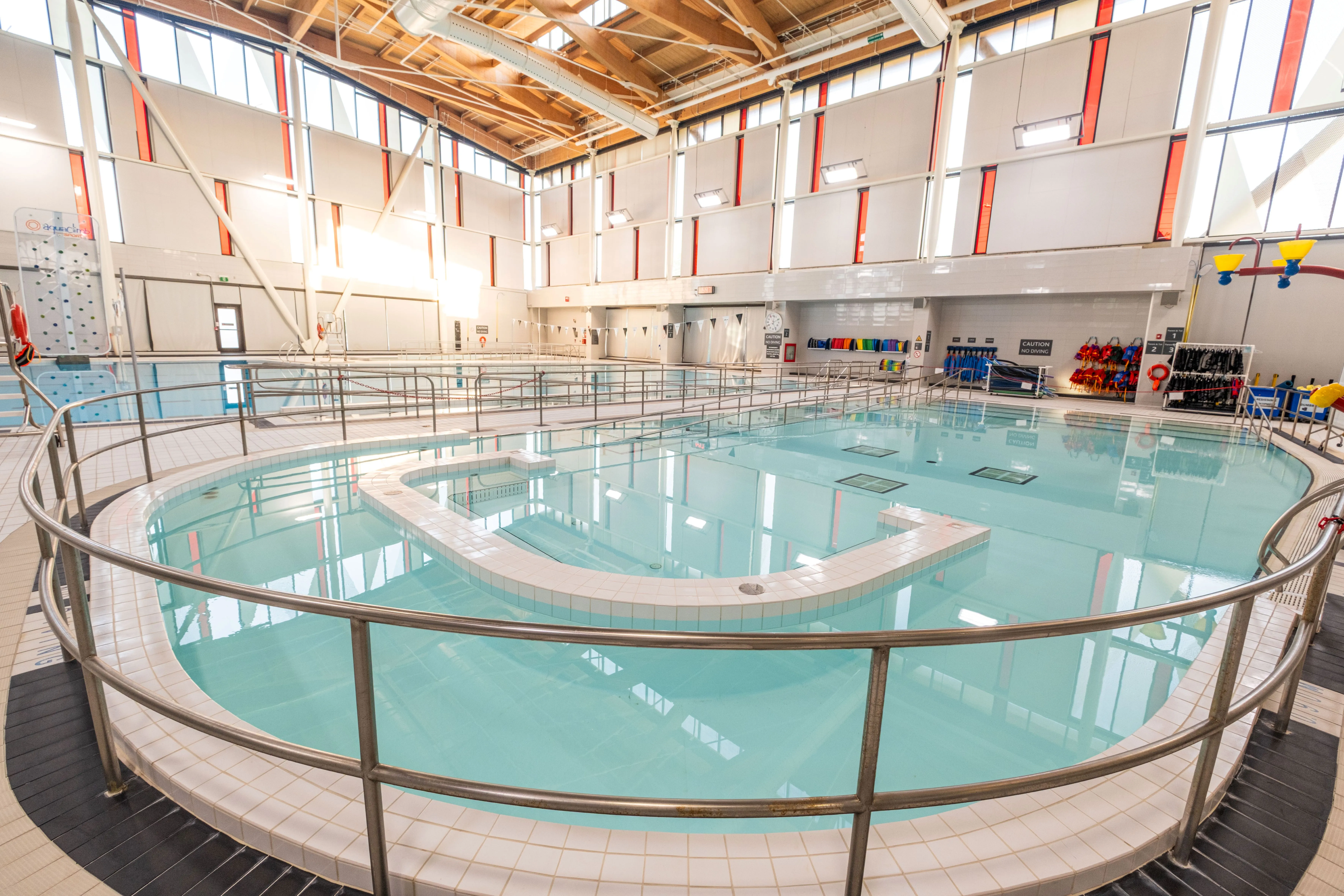 Indoor pool with clear water surrounded by safety railings and accessible ramps under a high ceiling with natural light.