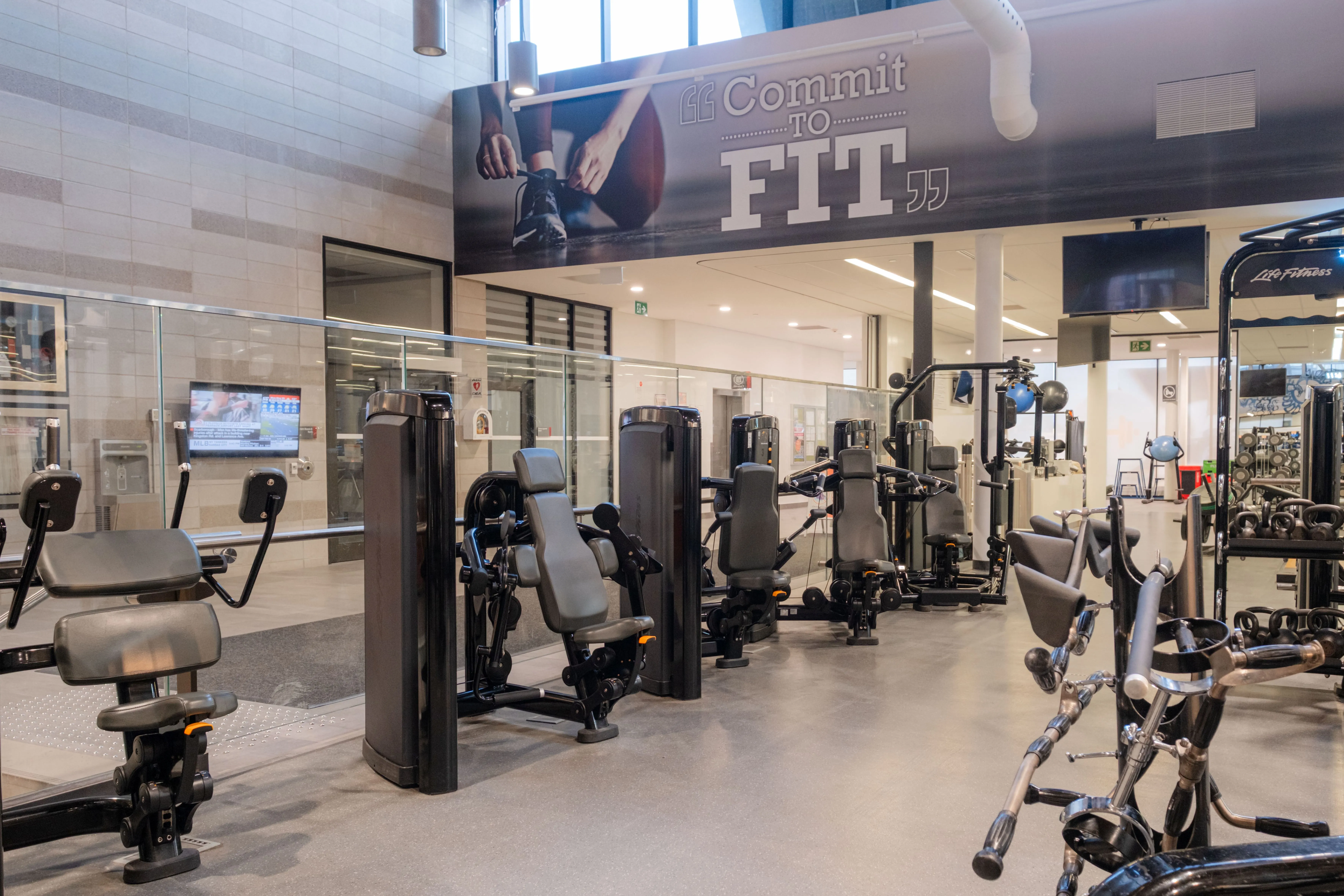 Modern gym interior with various strength training machines and a motivational 'Commit to Fit' banner overhead.