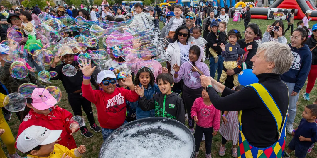 Children and families gather outdoors as a performer creates a stream of large soap bubbles, with many people reaching out to catch them at a community event.