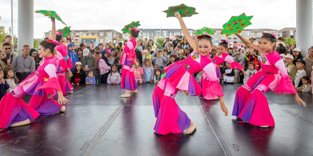 A group of performers in bright pink and purple costumes dance on an outdoor stage while holding decorative green fans, with a large crowd watching in the background.