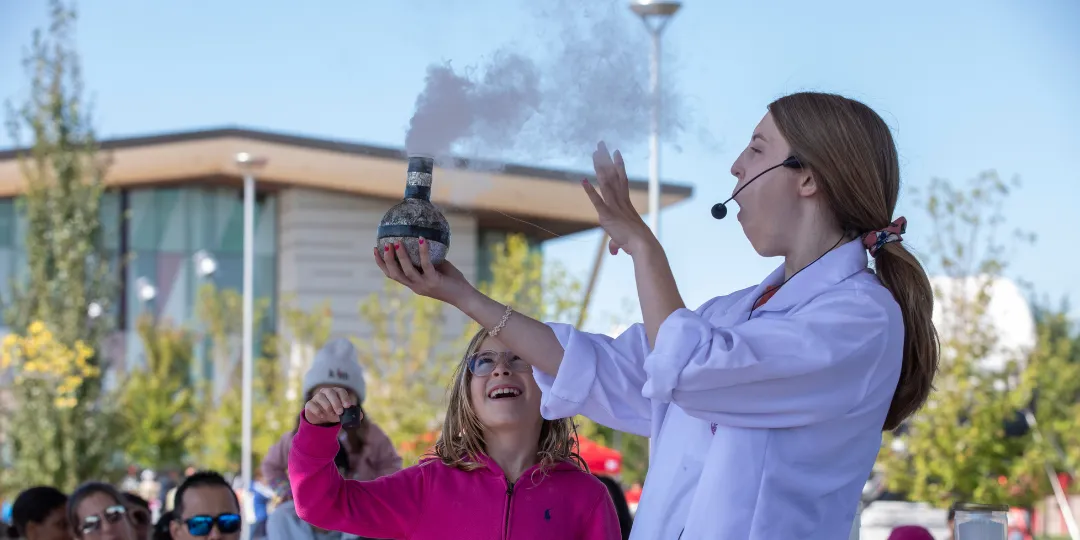 A science presenter wearing a lab coat demonstrates a smoking flask outdoors while a child in a pink jacket watches, with a crowd gathered in the background.