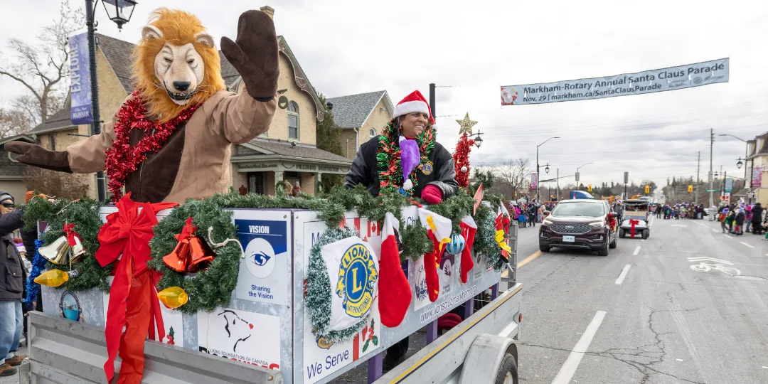 A festive parade float decorated with holiday wreaths, red bows and seasonal displays travels along a city street. characters stand on the float while spectators watch from the roadside.